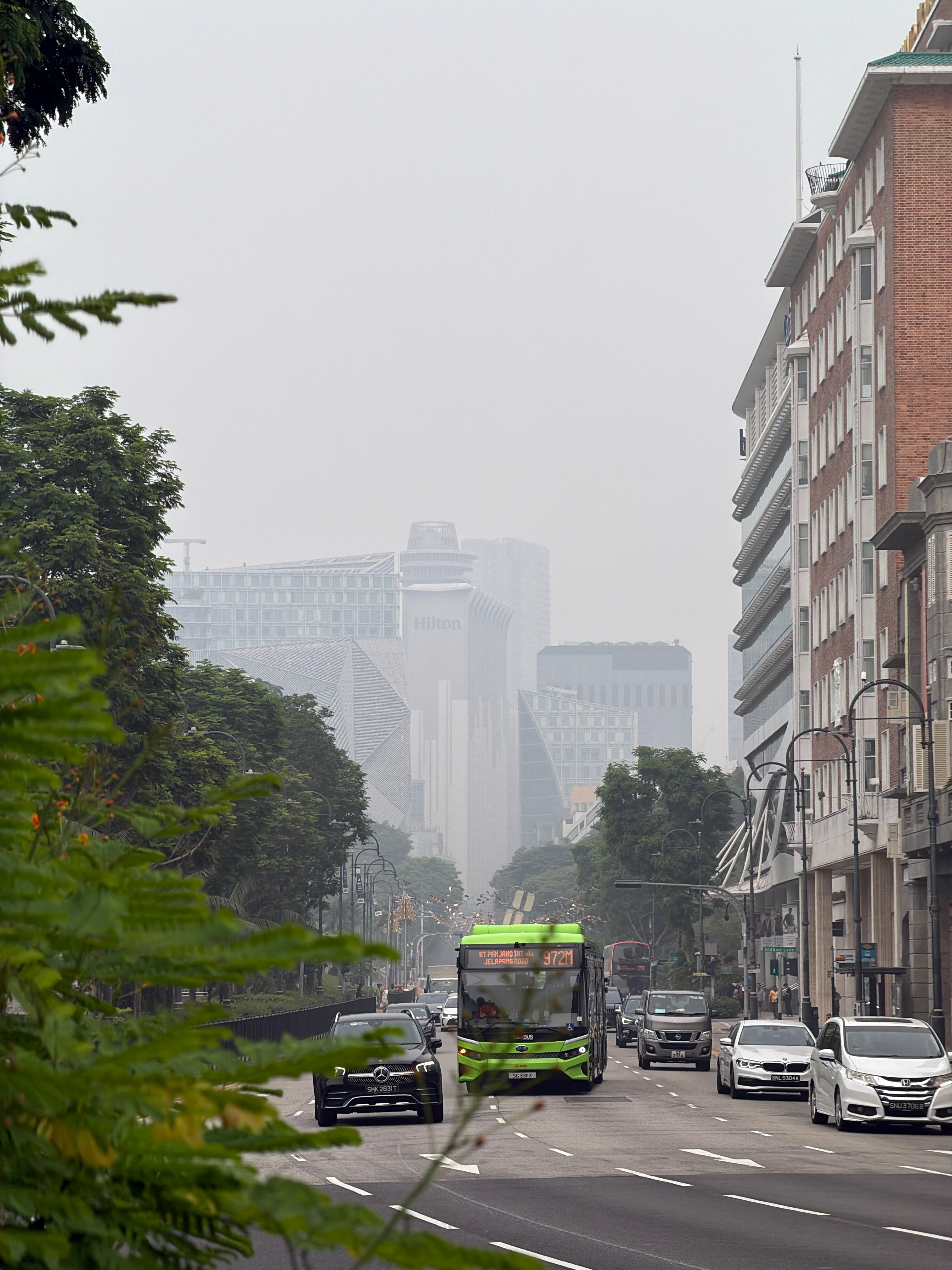 Traffic along Orchard Road in Singapore, with a green public bus and distant buildings partially obscured by thick haze.